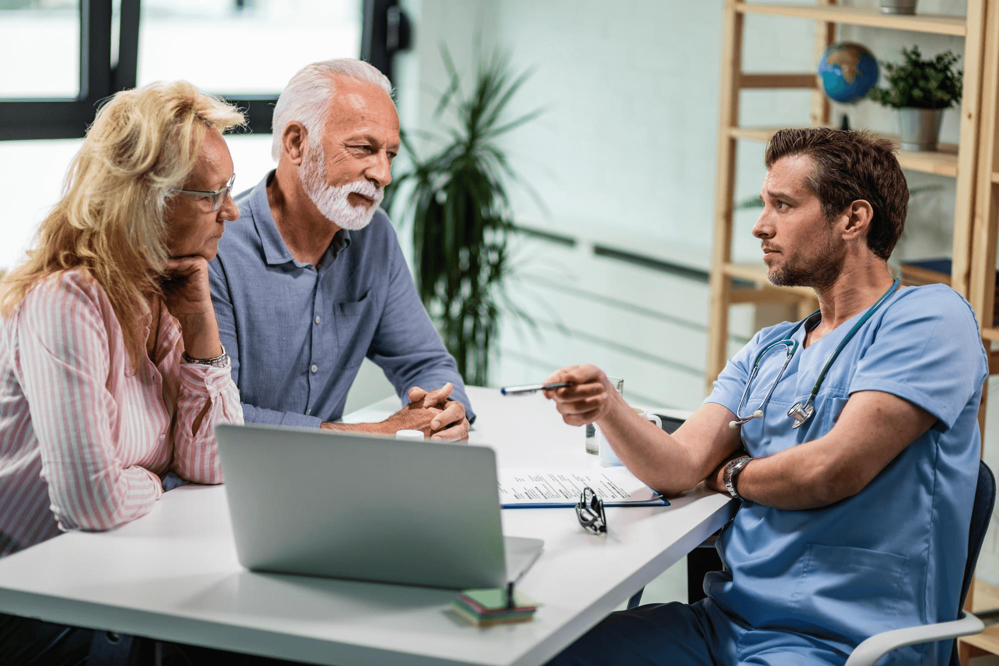 Physician in scrubs consulting with patients at a desk with a laptop and tablet